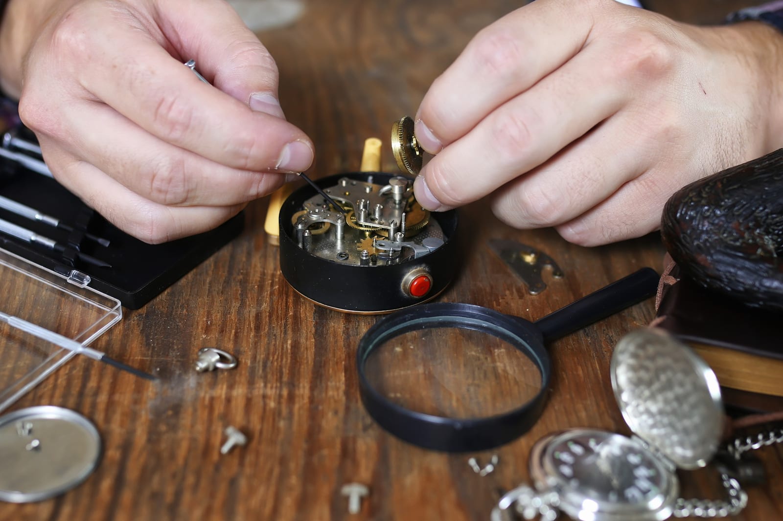 Due mani al lavoro su un movimento meccanico aperto, con lente d'ingrandimento e orologi da tasca sul banco